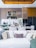 A contemporary kitchen and living area featuring a modern dining table set with wine glasses, plates, and cutlery. The kitchen is equipped with sleek cabinetry, a refrigerator, and decorative items like bottles and bowls. Above the dining area, stylish lighting fixtures can be seen. A white sofa with numerous decorative cushions in various textures is in the foreground, while tall white flowers add an elegant touch.