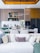 A contemporary kitchen and living area featuring a modern dining table set with wine glasses, plates, and cutlery. The kitchen is equipped with sleek cabinetry, a refrigerator, and decorative items like bottles and bowls. Above the dining area, stylish lighting fixtures can be seen. A white sofa with numerous decorative cushions in various textures is in the foreground, while tall white flowers add an elegant touch.