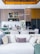 A contemporary kitchen and living area featuring a modern dining table set with wine glasses, plates, and cutlery. The kitchen is equipped with sleek cabinetry, a refrigerator, and decorative items like bottles and bowls. Above the dining area, stylish lighting fixtures can be seen. A white sofa with numerous decorative cushions in various textures is in the foreground, while tall white flowers add an elegant touch.