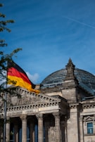 A historical building with classical architecture featuring a large glass dome on top and ornate stone carvings. A German flag with black, red, and yellow colors waves in the wind on a flagpole in the foreground. The sky is clear blue with a few wisps of clouds and a vapor trail from an airplane.