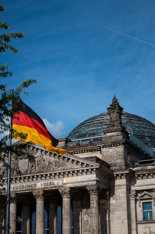 A historical building with classical architecture featuring a large glass dome on top and ornate stone carvings. A German flag with black, red, and yellow colors waves in the wind on a flagpole in the foreground. The sky is clear blue with a few wisps of clouds and a vapor trail from an airplane.