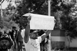 A black and white photograph of a protest where a person is holding up a sign that reads 'Buhari must go'. Several people are in the background, some obscured by trees, and there seems to be an intense atmosphere.