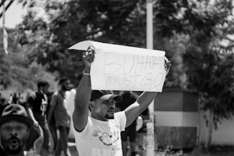 A black and white photograph of a protest where a person is holding up a sign that reads 'Buhari must go'. Several people are in the background, some obscured by trees, and there seems to be an intense atmosphere.