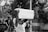 A black and white photograph of a protest where a person is holding up a sign that reads 'Buhari must go'. Several people are in the background, some obscured by trees, and there seems to be an intense atmosphere.