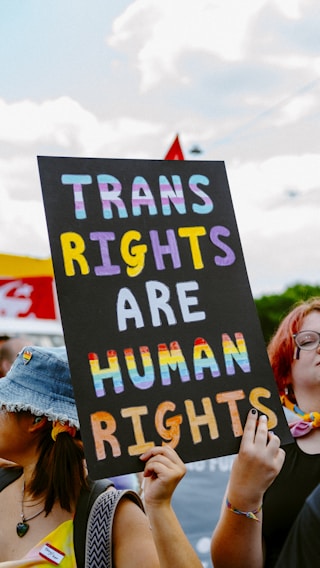 A protest with participants holding a sign advocating for transgender rights. The sign has bold lettering with the phrase 'TRANS RIGHTS ARE HUMAN RIGHTS' prominently displayed. The letters are colored with a rainbow gradient, symbolizing inclusivity and pride.