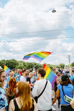 A vibrant outdoor gathering of people celebrating with rainbow flags. The scene is lively, with participants dressed in colorful attire. The background features green trees and a cloudy sky, adding to the festive atmosphere.