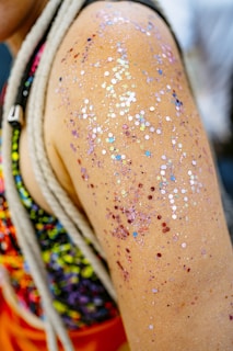 A close-up of a person's shoulder adorned with multicolored glitter. The skin is decorated with an array of shiny, reflective particles in various colors such as red, blue, silver, and pink. The background is partially visible, showing hints of a colorful garment.