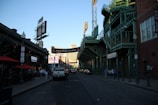 A street scene outside a stadium known as Fenway Park, with its iconic green structure visible. The street is lined with buildings and advertisements, including a cannabis dispensary. People are walking along the sidewalk while vehicles are seen on the road. A clear sky and setting sun cast shadows.