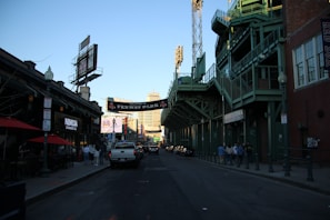 A street scene outside a stadium known as Fenway Park, with its iconic green structure visible. The street is lined with buildings and advertisements, including a cannabis dispensary. People are walking along the sidewalk while vehicles are seen on the road. A clear sky and setting sun cast shadows.