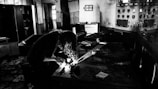 A dimly lit room with old control panels and scattered papers. A person with curly hair is bent over a table, using a flashlight, possibly examining objects or documents. The atmosphere is gritty and abandoned, with signs of neglect and decay.