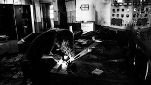A dimly lit room with old control panels and scattered papers. A person with curly hair is bent over a table, using a flashlight, possibly examining objects or documents. The atmosphere is gritty and abandoned, with signs of neglect and decay.