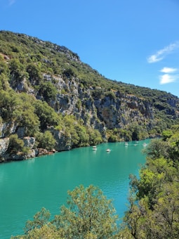 A scenic landscape featuring a vibrant turquoise river winding through lush green cliffs. Several small boats are gently floating on the water. The sky is clear with a few wispy clouds.