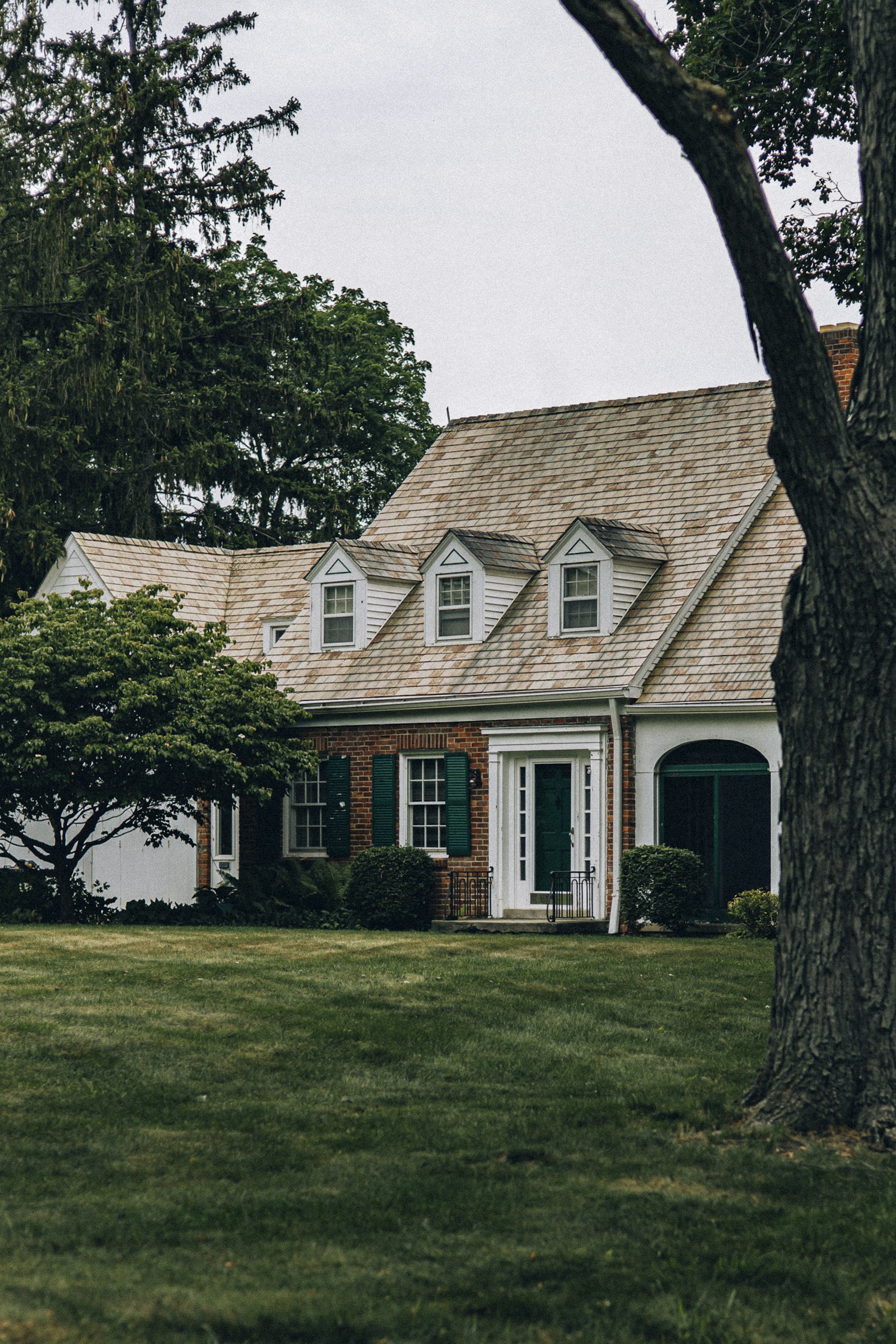 Brick traditional with a cedar-shingle roof, three dormers and green shutters behind a mature tree