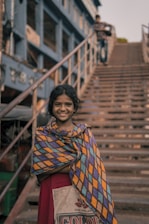 A young girl stands in front of a staircase wearing a colorful, patterned shawl over a red dress. She smiles warmly at the camera. In the background, a person is seen walking up the stairs. The setting appears to be urban with a teal-colored building along the side of the staircase.