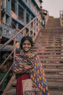 A young girl stands in front of a staircase wearing a colorful, patterned shawl over a red dress. She smiles warmly at the camera. In the background, a person is seen walking up the stairs. The setting appears to be urban with a teal-colored building along the side of the staircase.