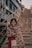 A young girl stands in front of a staircase wearing a colorful, patterned shawl over a red dress. She smiles warmly at the camera. In the background, a person is seen walking up the stairs. The setting appears to be urban with a teal-colored building along the side of the staircase.