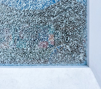 A large, rectangular section of cracked glass with intricate, web-like patterns spreading out from a focal point. The background is a mix of muted colors, creating an abstract and fragmented effect. The frame of the glass is made of a light-colored material, possibly concrete.