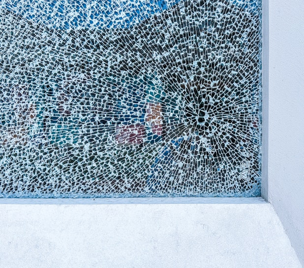 A large, rectangular section of cracked glass with intricate, web-like patterns spreading out from a focal point. The background is a mix of muted colors, creating an abstract and fragmented effect. The frame of the glass is made of a light-colored material, possibly concrete.