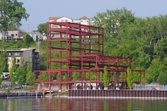 A red steel building framework stands prominently on the water's edge, surrounded by lush green trees and residential buildings in the background. The structure appears incomplete, with no walls or roof, and is set against a clear blue sky. The water in the foreground reflects the building and nearby greenery.