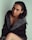 A woman with dark hair poses in a stylish coat with a fur trim. The setting is minimalistic with a soft, light background that contrasts with the dark clothing. She is making direct eye contact with the camera, and her expression is calm and confident.