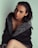 A woman with dark hair poses in a stylish coat with a fur trim. The setting is minimalistic with a soft, light background that contrasts with the dark clothing. She is making direct eye contact with the camera, and her expression is calm and confident.