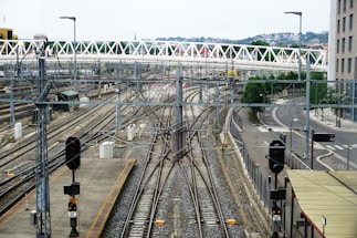 A railway yard with multiple train tracks converging and diverging. Overhead wires and signals are visible, indicating a complex rail infrastructure. A modern pedestrian bridge spans across the tracks, and urban buildings and roads surround the area. The environment appears orderly and structured with distant views of a hill and residential buildings in the background.