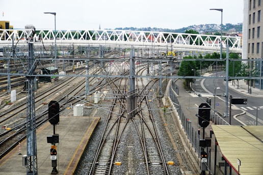A railway yard with multiple train tracks converging and diverging. Overhead wires and signals are visible, indicating a complex rail infrastructure. A modern pedestrian bridge spans across the tracks, and urban buildings and roads surround the area. The environment appears orderly and structured with distant views of a hill and residential buildings in the background.
