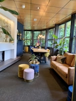 A cozy and modern interior space featuring a long wooden dining table surrounded by colorful chairs. The room is well-lit by multiple pendant lights hanging from the ceiling, and large windows provide a view of lush greenery outside. There are several potted plants adding a touch of nature indoors. A comfortable brown sofa is paired with soft, colorful poufs, and a small table with more plants adds to the decor. The ceiling has a warm wooden finish that complements the overall earthy tones of the room.