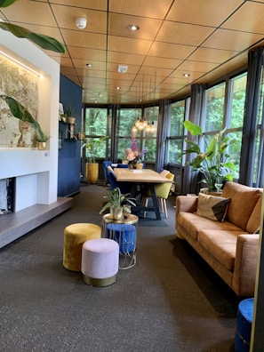 A cozy and modern interior space featuring a long wooden dining table surrounded by colorful chairs. The room is well-lit by multiple pendant lights hanging from the ceiling, and large windows provide a view of lush greenery outside. There are several potted plants adding a touch of nature indoors. A comfortable brown sofa is paired with soft, colorful poufs, and a small table with more plants adds to the decor. The ceiling has a warm wooden finish that complements the overall earthy tones of the room.