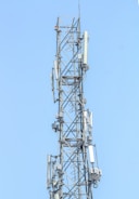 A telecommunications tower under construction with workers installing antenna equipment against a clear blue sky