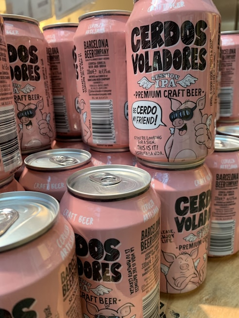 A collection of pink craft beer cans labeled 'Cerdos Voladores' from Barcelona Beer Company. The cans feature cartoon images of a pig wearing sunglasses with angel wings, along with various textual and design elements. The cans are arranged closely together on a wooden surface.