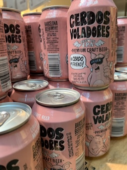 A collection of pink craft beer cans labeled 'Cerdos Voladores' from Barcelona Beer Company. The cans feature cartoon images of a pig wearing sunglasses with angel wings, along with various textual and design elements. The cans are arranged closely together on a wooden surface.