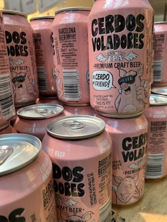 A collection of pink craft beer cans labeled 'Cerdos Voladores' from Barcelona Beer Company. The cans feature cartoon images of a pig wearing sunglasses with angel wings, along with various textual and design elements. The cans are arranged closely together on a wooden surface.