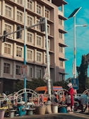A bustling street scene featuring a market setting in front of a multi-story building with numerous windows. People, mostly women, are engaging in selling goods like onions displayed in baskets. A pedestrian crossing sign and traffic lights are present, with a colorful bus parked alongside.