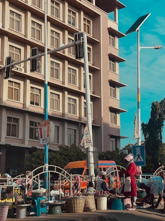 A bustling street scene featuring a market setting in front of a multi-story building with numerous windows. People, mostly women, are engaging in selling goods like onions displayed in baskets. A pedestrian crossing sign and traffic lights are present, with a colorful bus parked alongside.