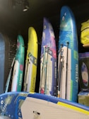 Several colorful stand-up paddleboards are displayed vertically against a dark wall. The boards have diverse designs, featuring blue, yellow, green, and pink hues, with the brand name 'Aztron' visible on each. The modern setup includes additional accessories like paddles and a bag on the side.