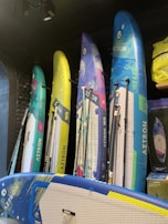 Several colorful stand-up paddleboards are displayed vertically against a dark wall. The boards have diverse designs, featuring blue, yellow, green, and pink hues, with the brand name 'Aztron' visible on each. The modern setup includes additional accessories like paddles and a bag on the side.