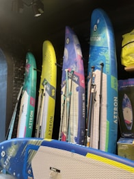 Several colorful stand-up paddleboards are displayed vertically against a dark wall. The boards have diverse designs, featuring blue, yellow, green, and pink hues, with the brand name 'Aztron' visible on each. The modern setup includes additional accessories like paddles and a bag on the side.