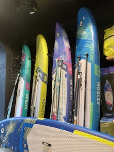 Several colorful stand-up paddleboards are displayed vertically against a dark wall. The boards have diverse designs, featuring blue, yellow, green, and pink hues, with the brand name 'Aztron' visible on each. The modern setup includes additional accessories like paddles and a bag on the side.