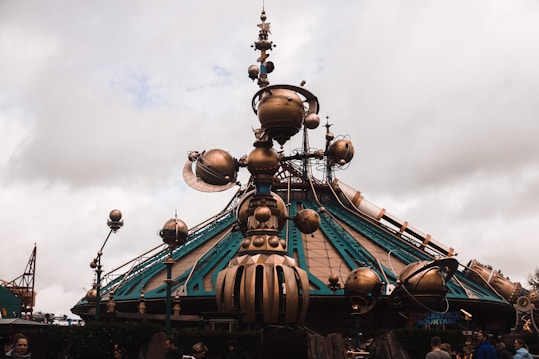A futuristic structure features a large dome-like base with intricate metallic decorations and globes extending outward. The design incorporates a blend of teal and bronze colors with mechanical and astronomical details. People are visible in the foreground, suggesting a theme park or exhibition setting.