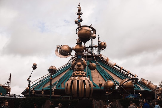 A futuristic structure features a large dome-like base with intricate metallic decorations and globes extending outward. The design incorporates a blend of teal and bronze colors with mechanical and astronomical details. People are visible in the foreground, suggesting a theme park or exhibition setting.