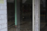 A view through a glass door shows an empty interior space with bare walls and flooring. The room appears to be under construction or renovation, with visible wiring and some debris on the ground. The glass reflects dim external lighting, and a faint outline of a car outside can be seen.