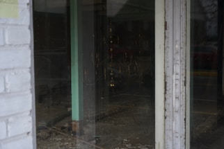 A view through a glass door shows an empty interior space with bare walls and flooring. The room appears to be under construction or renovation, with visible wiring and some debris on the ground. The glass reflects dim external lighting, and a faint outline of a car outside can be seen.
