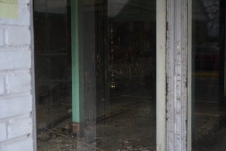 A view through a glass door shows an empty interior space with bare walls and flooring. The room appears to be under construction or renovation, with visible wiring and some debris on the ground. The glass reflects dim external lighting, and a faint outline of a car outside can be seen.