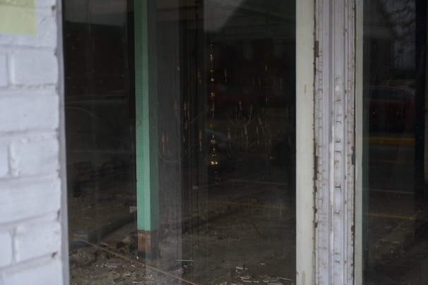 A view through a glass door shows an empty interior space with bare walls and flooring. The room appears to be under construction or renovation, with visible wiring and some debris on the ground. The glass reflects dim external lighting, and a faint outline of a car outside can be seen.