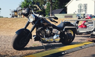 A black motorcycle with a shiny chrome engine is parked in an outdoor area, with a helmet placed on its handlebars. The motorcycle is positioned near a sidewalk, and in the background are a residential building, some trees, and a fence. Next to the motorcycle, there is a red scooter secured to a bike rack.