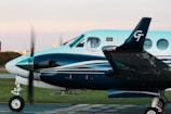 A turquoise and white twin-engine propeller aircraft with a person visible in the cockpit, parked on a tarmac. The Brazilian flag and an airline logo are displayed near the cockpit window, with blurred propellers indicating slight motion. In the background, there is a hint of an urban landscape and a green grass field.