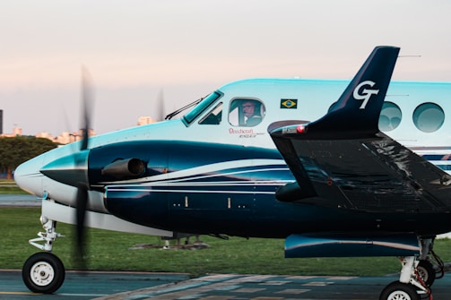 A turquoise and white twin-engine propeller aircraft with a person visible in the cockpit, parked on a tarmac. The Brazilian flag and an airline logo are displayed near the cockpit window, with blurred propellers indicating slight motion. In the background, there is a hint of an urban landscape and a green grass field.