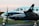 A turquoise and white twin-engine propeller aircraft with a person visible in the cockpit, parked on a tarmac. The Brazilian flag and an airline logo are displayed near the cockpit window, with blurred propellers indicating slight motion. In the background, there is a hint of an urban landscape and a green grass field.