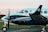 A turquoise and white twin-engine propeller aircraft with a person visible in the cockpit, parked on a tarmac. The Brazilian flag and an airline logo are displayed near the cockpit window, with blurred propellers indicating slight motion. In the background, there is a hint of an urban landscape and a green grass field.